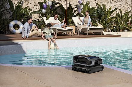 Robot limpiapiscinas Beatbot sobre el borde de una piscina exterior mientras una familia disfruta del sol en tumbonas al fondo