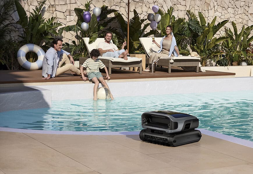 Robot limpiapiscinas Beatbot sobre el borde de una piscina exterior mientras una familia disfruta del sol en tumbonas al fondo