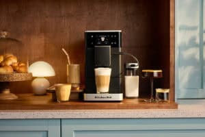 Cafetera espresso superautomática KitchenAid en negro sobre encimera de madera rodeada de tazas con latte macchiato, café con hielo y espresso junto a croissants y jarra de leche