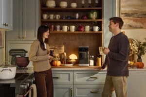 Pareja conversando en cocina moderna mientras sostienen bebidas de café preparadas con cafetera espresso superautomática KitchenAid sobre encimera con estantes de madera al fondo
