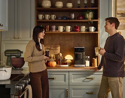 Pareja conversando en cocina moderna mientras sostienen bebidas de café preparadas con cafetera espresso superautomática KitchenAid sobre encimera con estantes de madera al fondo