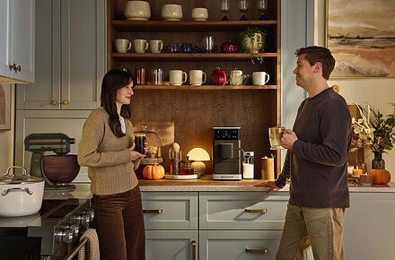 Pareja conversando en cocina moderna mientras sostienen bebidas de café preparadas con cafetera espresso superautomática KitchenAid sobre encimera con estantes de madera al fondo