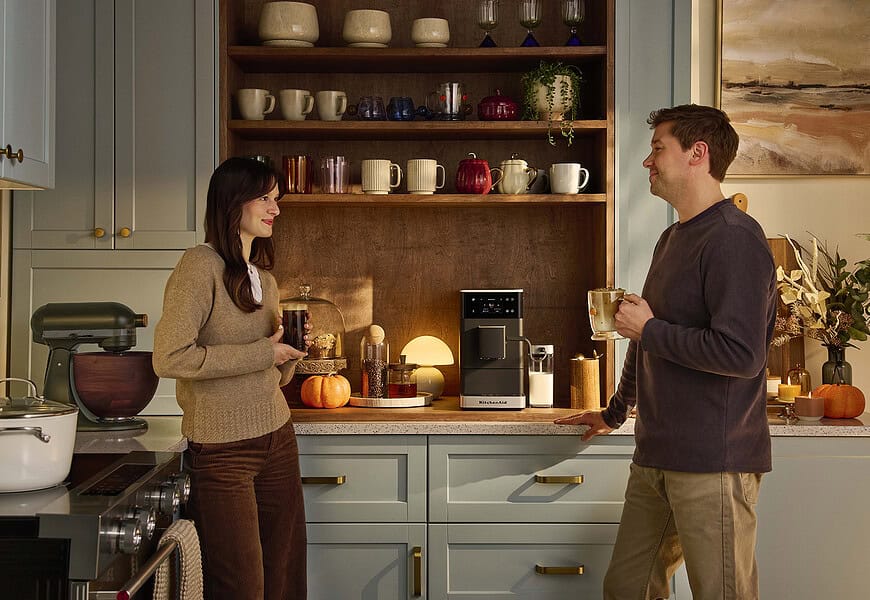 Pareja conversando en cocina moderna mientras sostienen bebidas de café preparadas con cafetera espresso superautomática KitchenAid sobre encimera con estantes de madera al fondo