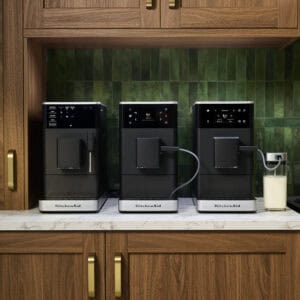 Tres modelos de cafeteras espresso superautomáticas KitchenAid en color negro alineadas sobre encimera de mármol blanco en cocina con armarios de madera oscura y azulejos verdes