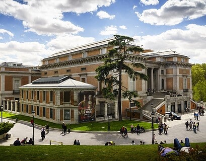 Vista exterior del Museo Nacional del Prado en Madrid con visitantes paseando por los jardines y cartel de la exposición Georges de La Tour en fachada