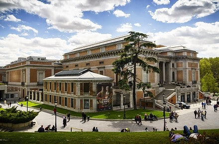 Vista exterior del Museo Nacional del Prado en Madrid con visitantes paseando por los jardines y cartel de la exposición Georges de La Tour en fachada