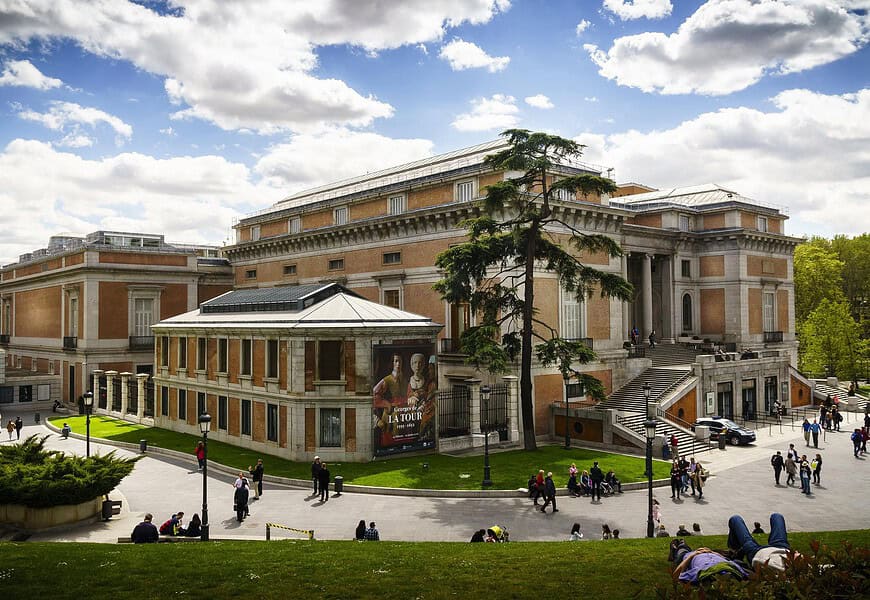 Vista exterior del Museo Nacional del Prado en Madrid con visitantes paseando por los jardines y cartel de la exposición Georges de La Tour en fachada