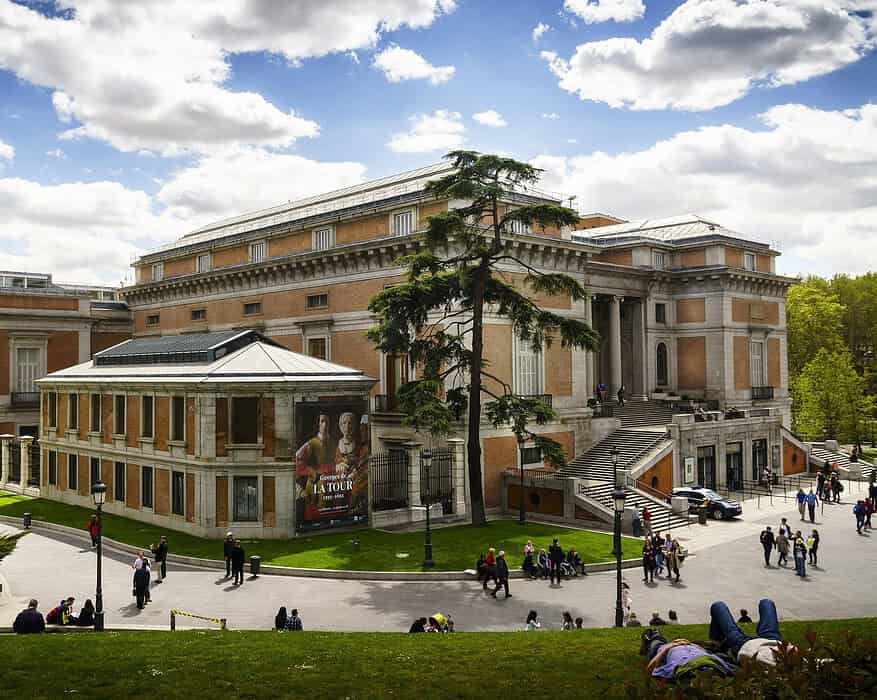 Vista exterior del Museo Nacional del Prado en Madrid con visitantes paseando por los jardines y cartel de la exposición Georges de La Tour en fachada