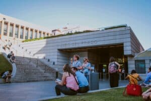 Visitantes sentados y de pie frente a la Puerta de los Jerónimos del Museo del Prado en Madrid al atardecer durante una jornada de puertas abiertas